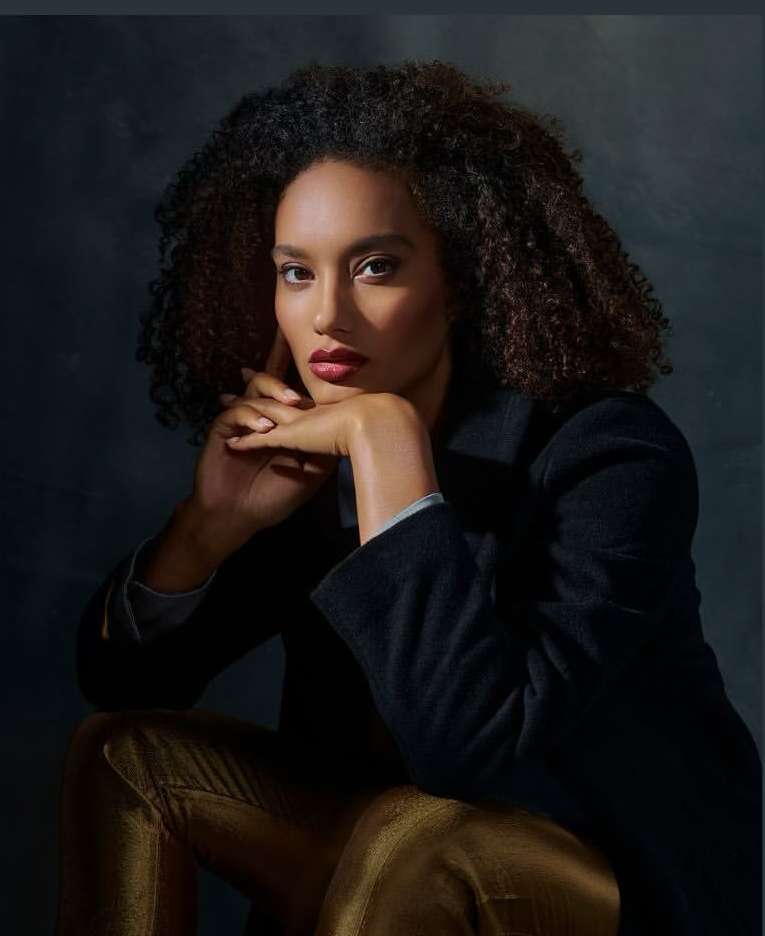 Portrait of a woman with curly hair in a black coat and gold pants, seated and posing in a studio with soft lighting.